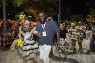 Tall bespectacled man in blue shirt being led in procession with Brazilian woman dancer in traditional colorful clothing