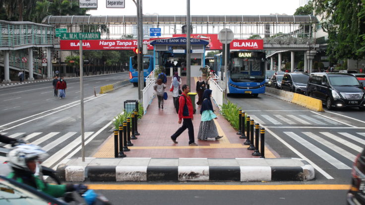 A picture of the Bank Indonesia Transjakarta BRT Station in Jakarta. People are walking on a crosswalk. A BRT bus is in the background in a BRT lane.
